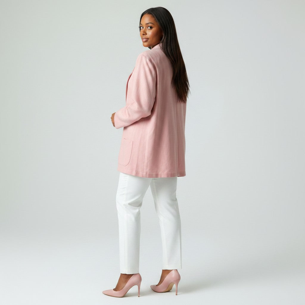 A woman stands in profile wearing a light pink blazer, white pants, and matching pink heels against a plain light gray studio background.