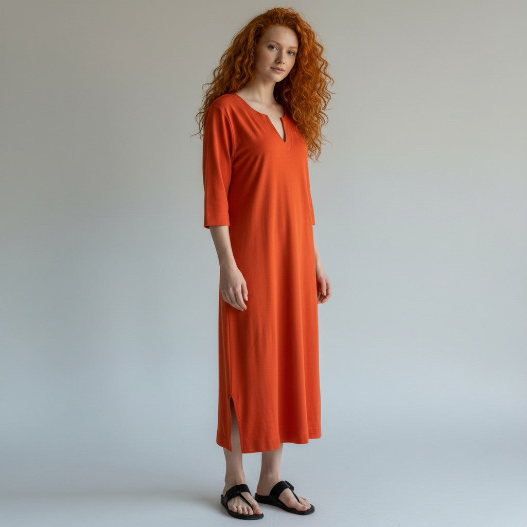 A woman with long curly red hair stands against a light gray studio background wearing a loose, knee-length orange dress with three-quarter sleeves and black sandals.
