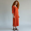 A woman with long curly red hair stands against a light gray studio background wearing a loose, knee-length orange dress with three-quarter sleeves and black sandals.