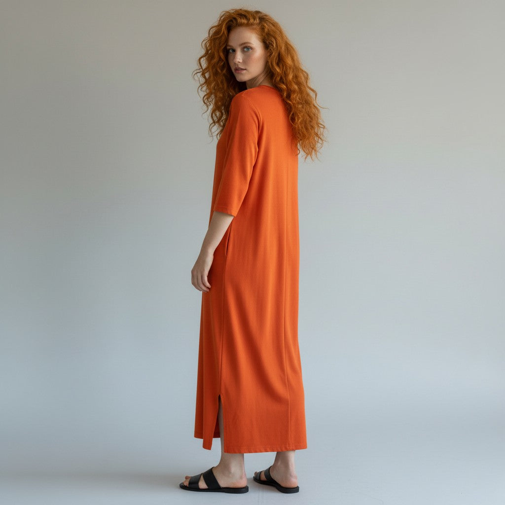 A woman with curly red hair wearing a bright orange midi dress with elbow-length sleeves and black slide sandals, photographed against a neutral light gray studio background.