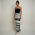A woman modeling a strapless black top paired with a long black and white striped skirt, standing against a plain light gray studio background.