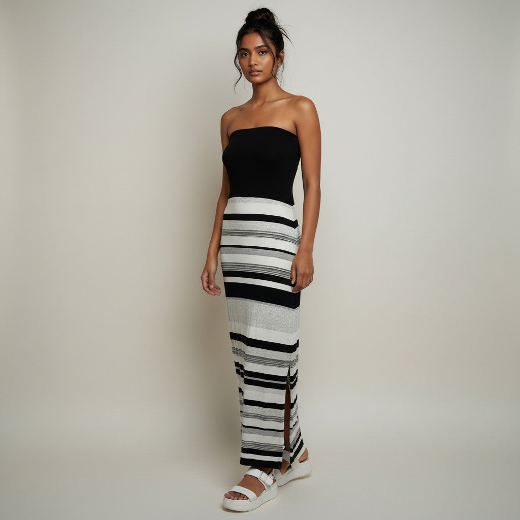 A woman modeling a strapless black top paired with a long black and white striped skirt, standing against a plain light gray studio background.