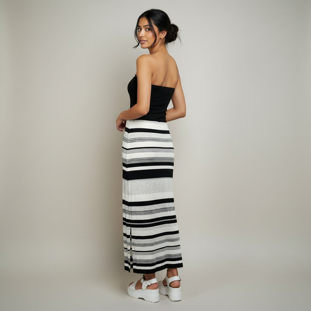 A woman stands in a studio wearing a black strapless top and a long black-and-white striped skirt with white platform sandals, photographed against a plain light gray background.