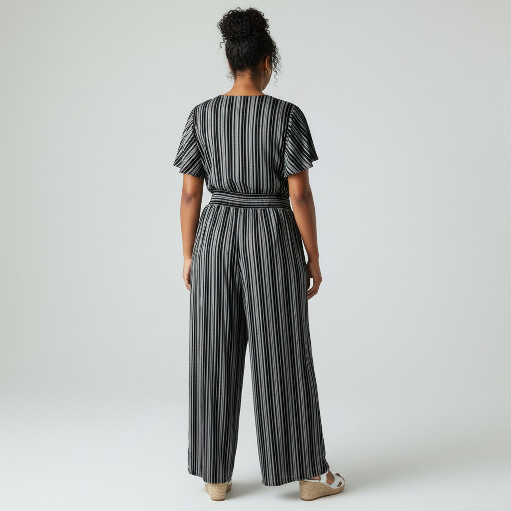 A woman viewed from the back wearing a black and white vertically striped short-sleeve jumpsuit with a matching waist belt, paired with beige wedge sandals, against a light gray studio background.