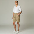 A model stands in a studio wearing a white short-sleeve button-up shirt, beige cargo shorts, and white sneakers against a light gray background.