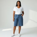 A woman modeling blue denim shorts with a white V-neck t-shirt and white sneakers, standing against a light gray studio background with soft diagonal shadows.