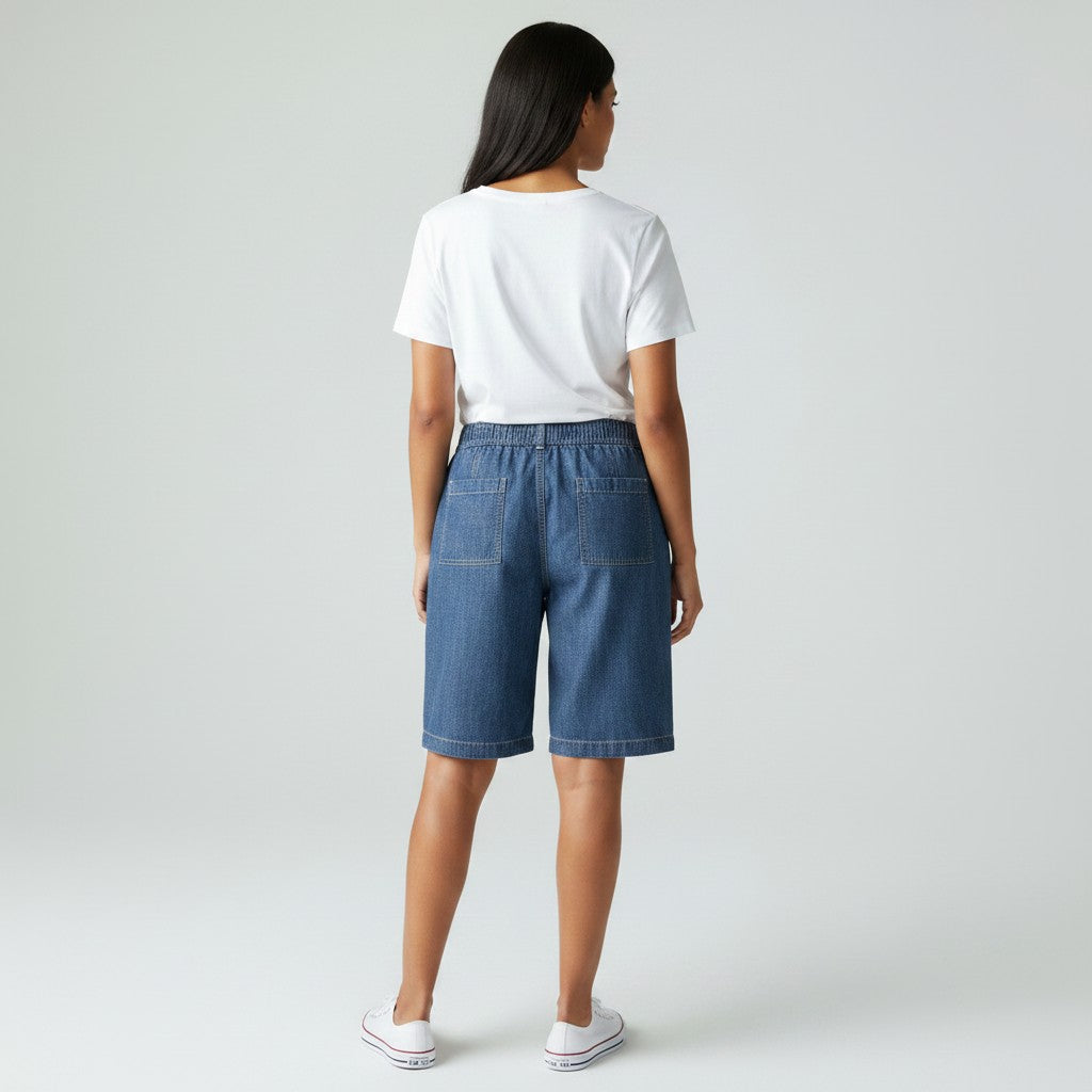 A woman viewed from behind wearing a white t-shirt, blue denim shorts, and white sneakers, standing against a light gray studio background.