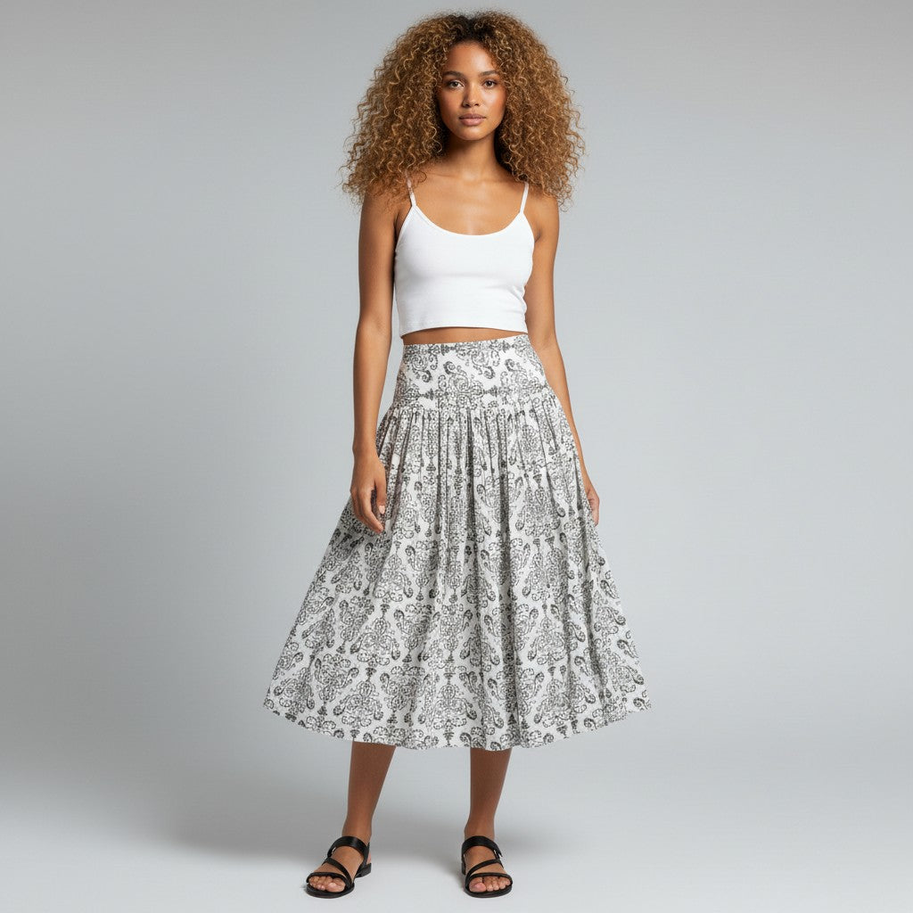 A woman stands against a light gray studio background wearing a white ribbed tank top and a high-waisted midi skirt with a black and white paisley pattern. She is wearing black strappy flat sandals.