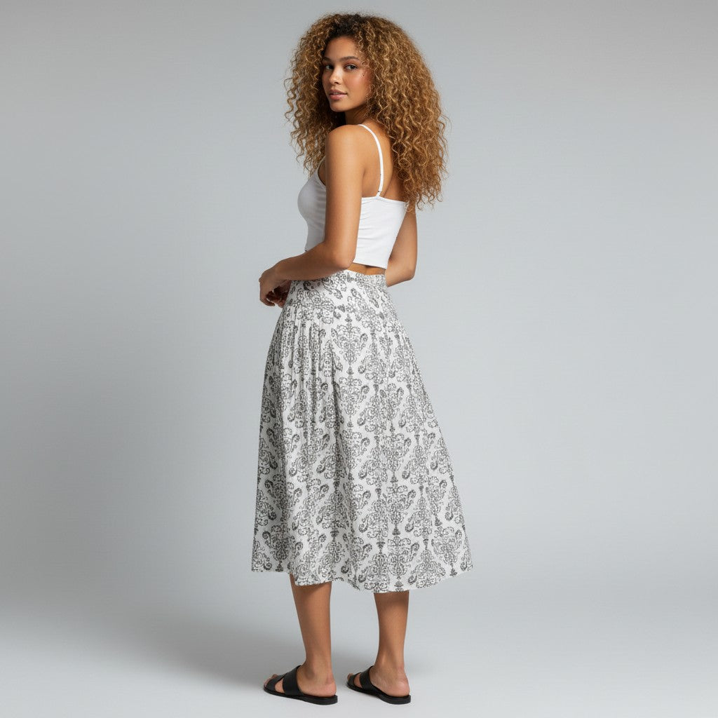 A woman with curly hair stands in profile wearing a white camisole top and a high-waisted midi skirt with a black and white paisley pattern, paired with black slide sandals, against a light gray studio background.
