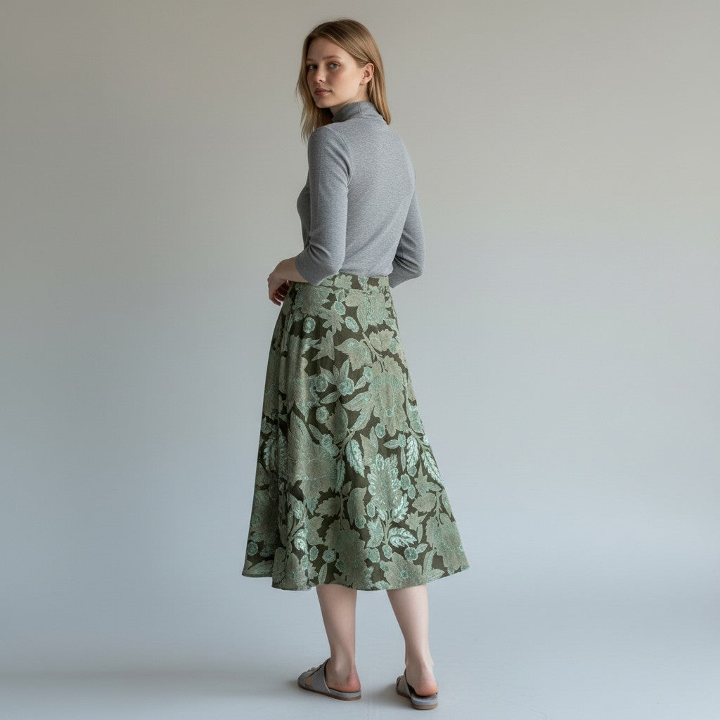 A woman stands in profile wearing a gray long-sleeve turtleneck top and a knee-length green floral skirt with a dark olive pattern, paired with gray slip-on shoes, against a plain light gray studio background.