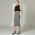 A woman modeling a sleeveless black collared top and a black-and-white horizontally striped knee-length pencil skirt, paired with black high heels, against a light gray studio background with soft window-like shadows.