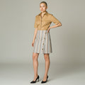 A model stands against a light gray studio background wearing a tan short-sleeve button-up shirt and a high-waisted A-line mini skirt with vertical stripes in beige, white, and brown, featuring decorative buttons down the front. She is wearing black pointed-toe heels.