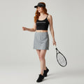 A woman modeling a gray athletic tennis skirt with a black sports bra and black visor, holding a tennis racket against a light gray studio background.