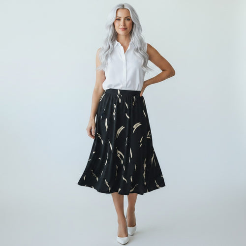 A woman with long silver hair models a sleeveless white button-up shirt and a black midi skirt with a beige feather pattern, paired with white pointed-toe heels, against a light gray studio background.
