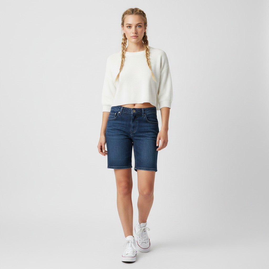 A woman modeling a white cropped long-sleeve knit top and dark blue denim shorts against a light gray studio background.