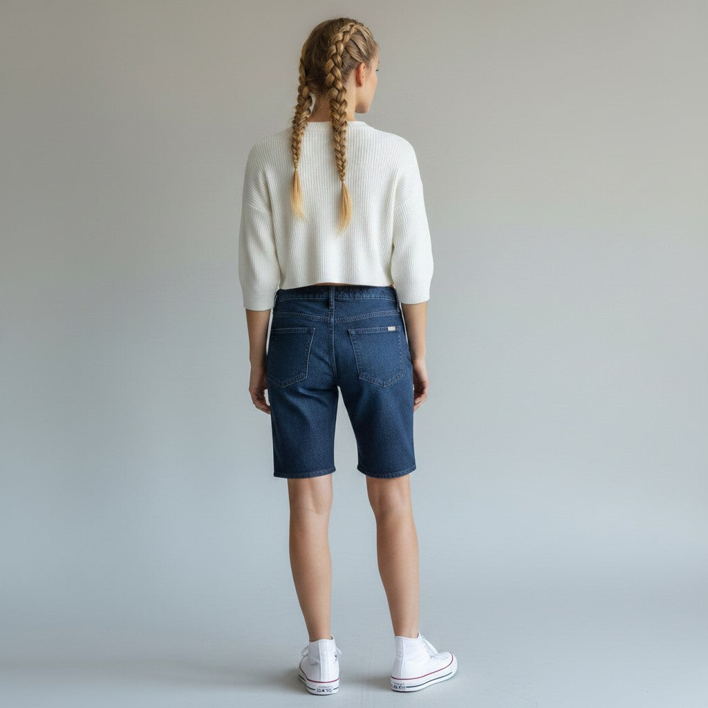 A child model shown from the back wearing a white knit cropped sweater, dark blue denim shorts, and white sneakers against a light gray studio background.