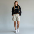 A model with long braided hair stands against a light gray studio background wearing a black cropped hoodie with white text reading 'Reclaimed Revival Revived,' light beige shorts, and white sneakers.