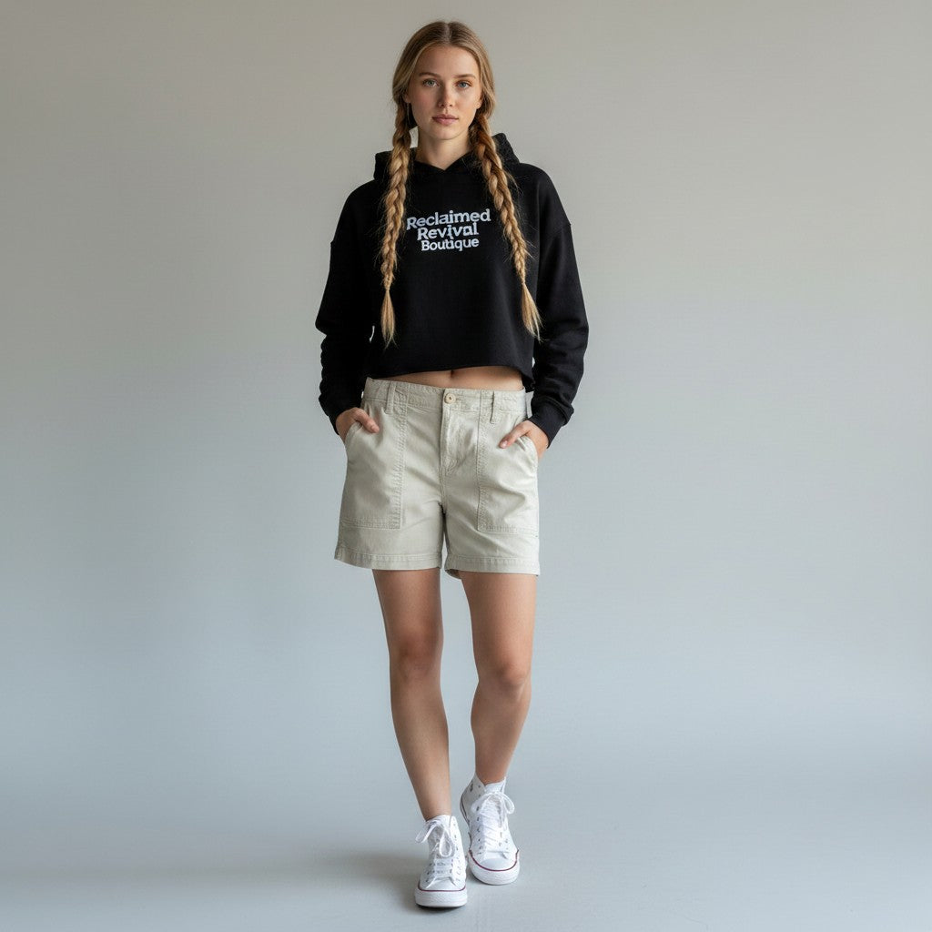 A model with long braided hair stands against a light gray studio background wearing a black cropped hoodie with white text reading 'Reclaimed Revival Revived,' light beige shorts, and white sneakers.