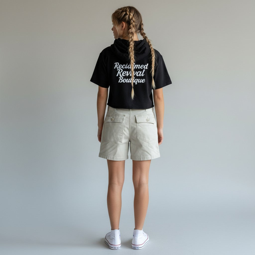 A girl with two braids is shown from the back wearing a black cropped t-shirt with white text that reads 'Reclaimed Revival Boutique' and beige cargo shorts, standing against a light gray studio background.