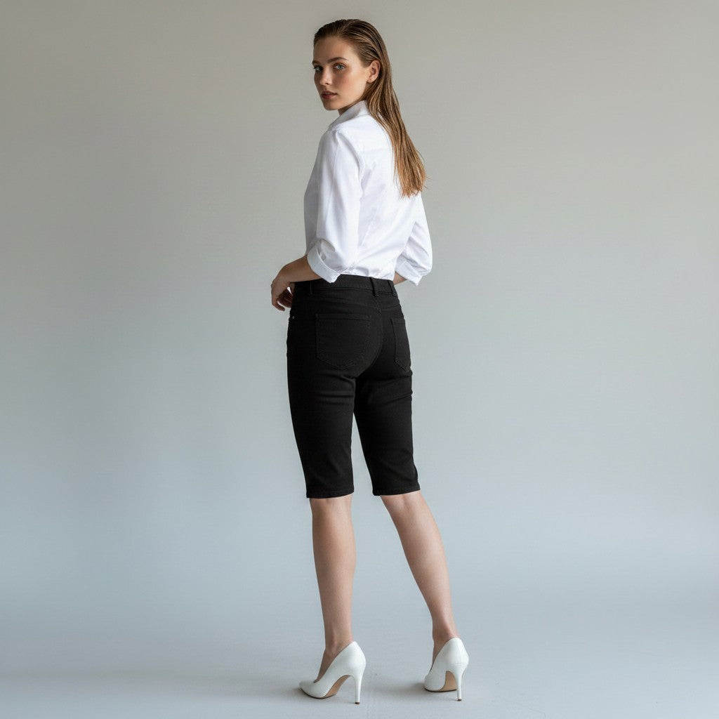 A woman posing from the back wearing black knee-length shorts, a white button-down shirt, and white high heels against a light gray studio background.