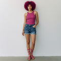A woman with a bright magenta afro stands against a plain white wall wearing a sleeveless magenta tank top, blue denim shorts, and matching pink strappy sandals.