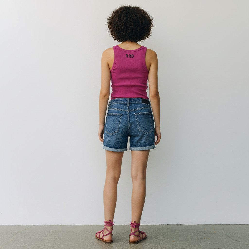 A woman is shown from behind wearing a bright pink ribbed tank top with a small black logo on the back, blue denim shorts, and pink strappy sandals, standing against a plain white wall.