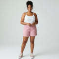 A woman modeling a white spaghetti-strap tank top and light pink denim shorts with distressed details, paired with colorful sneakers, against a light gray studio background.