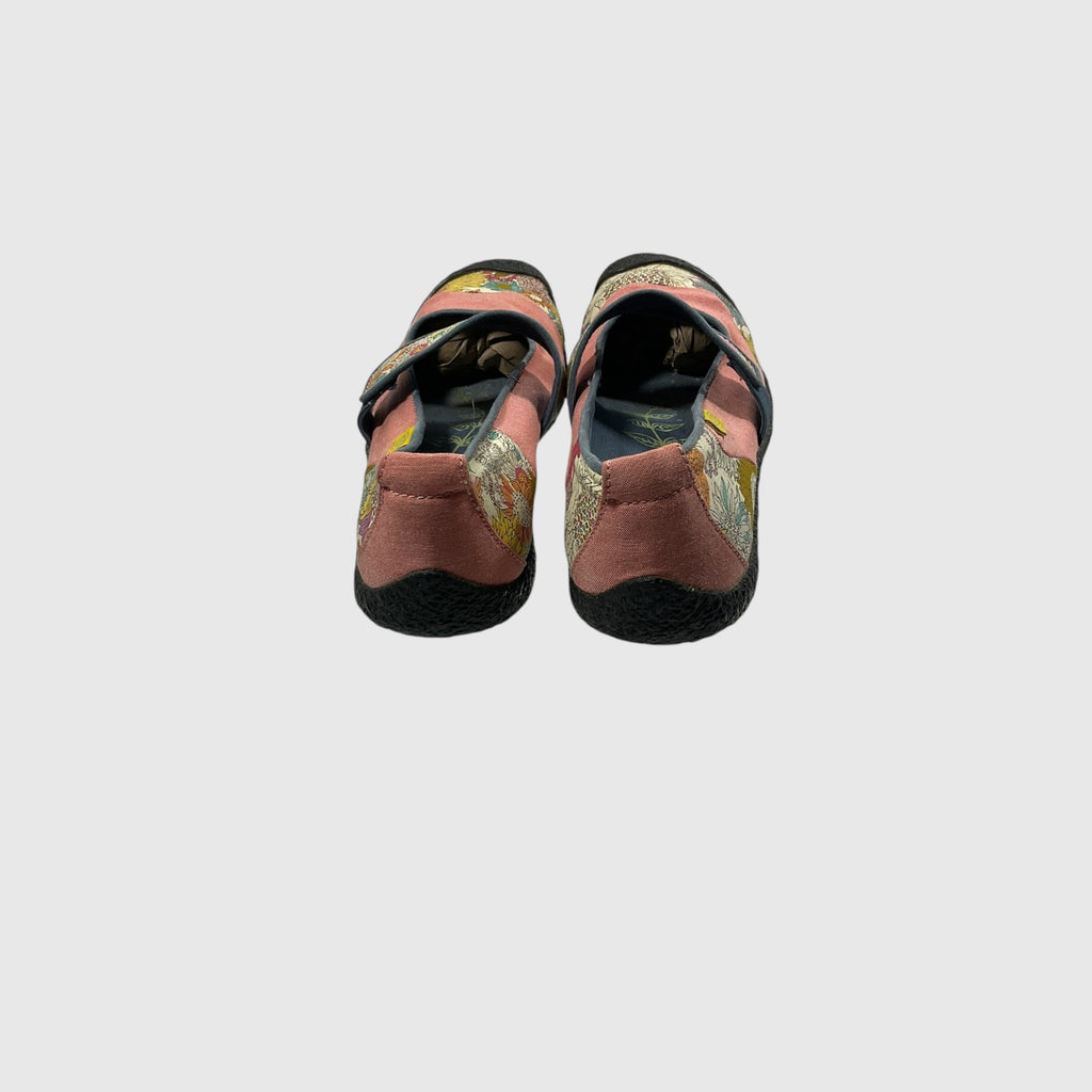 A pair of children's flat shoes shown from the back, featuring a dusty rose suede upper with a colorful floral pattern on the sides, black soles, and hook-and-loop strap closures, set against a solid light gray background.