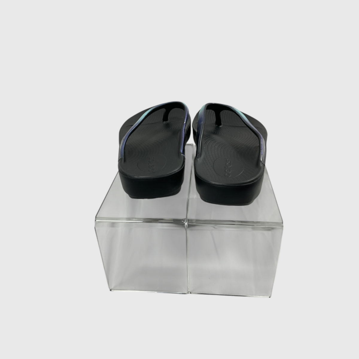 A pair of black flip-flop sandals with thin blue straps, displayed from the back on a clear acrylic block against a light gray background.