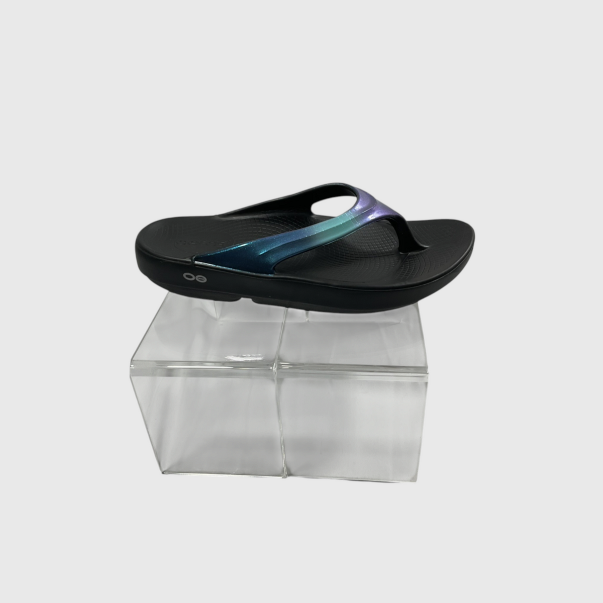 A black flip-flop sandal with a glossy iridescent strap featuring a blue-to-purple gradient, displayed on a clear acrylic block against a light gray background.