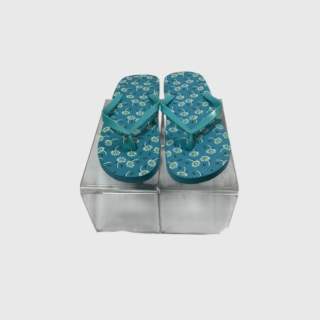 A pair of teal flip-flops with a white and green floral pattern, displayed on a clear plastic block against a light gray background.