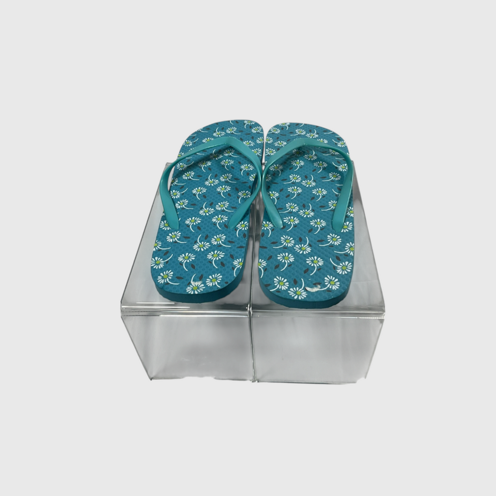 A pair of teal flip-flops with a white floral pattern, displayed on a clear acrylic block against a light gray background.