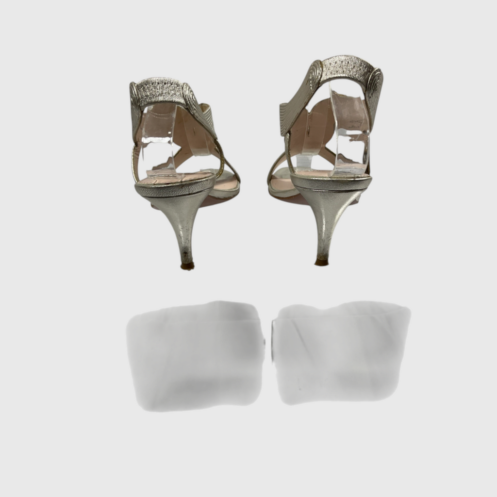 A pair of metallic silver high-heeled sandals with clear side panels and silver straps, shown from the back on a light gray background with a faint reflection below.