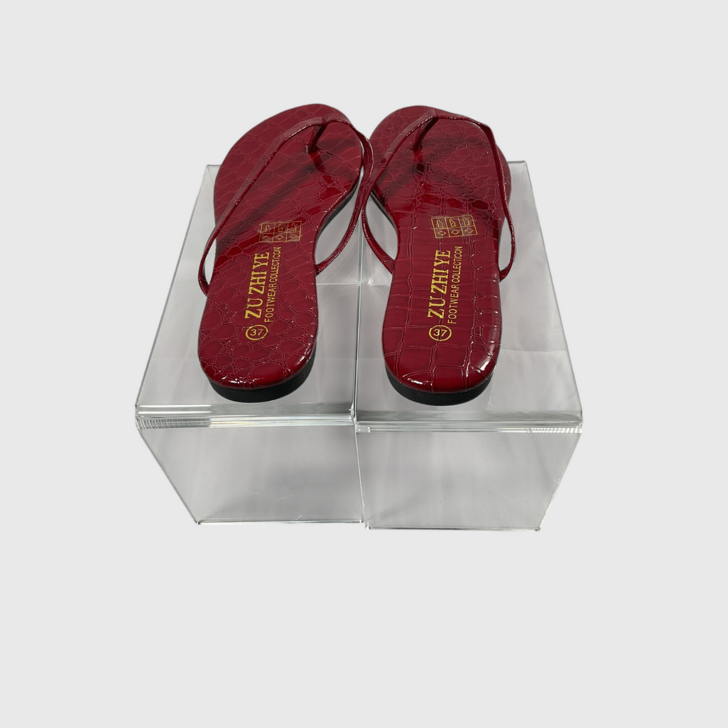 A pair of red crocodile-embossed thong sandals with gold 'ALZAHVE' branding on the soles, displayed on a clear acrylic block against a light gray background.