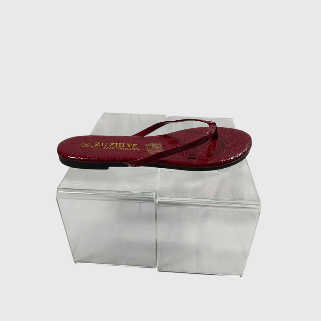 A single red crocodile-embossed thong sandal with gold branding on the sole, displayed on a clear acrylic block against a light gray background.