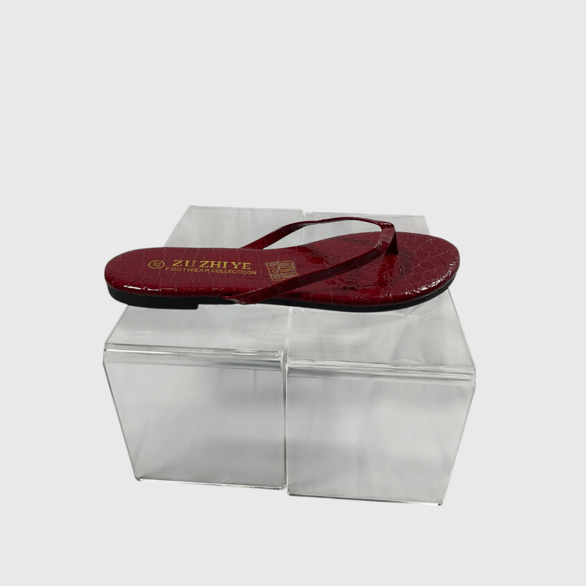 A single red crocodile-embossed thong sandal with gold branding on the sole, displayed on a clear acrylic block against a light gray background.