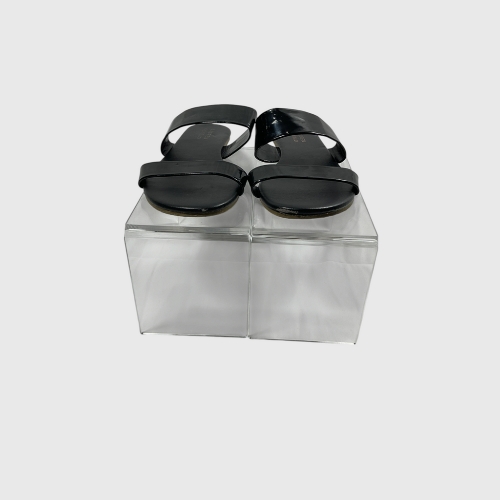 A pair of black patent leather slide sandals with wide double straps, displayed on a clear acrylic block against a light gray background.