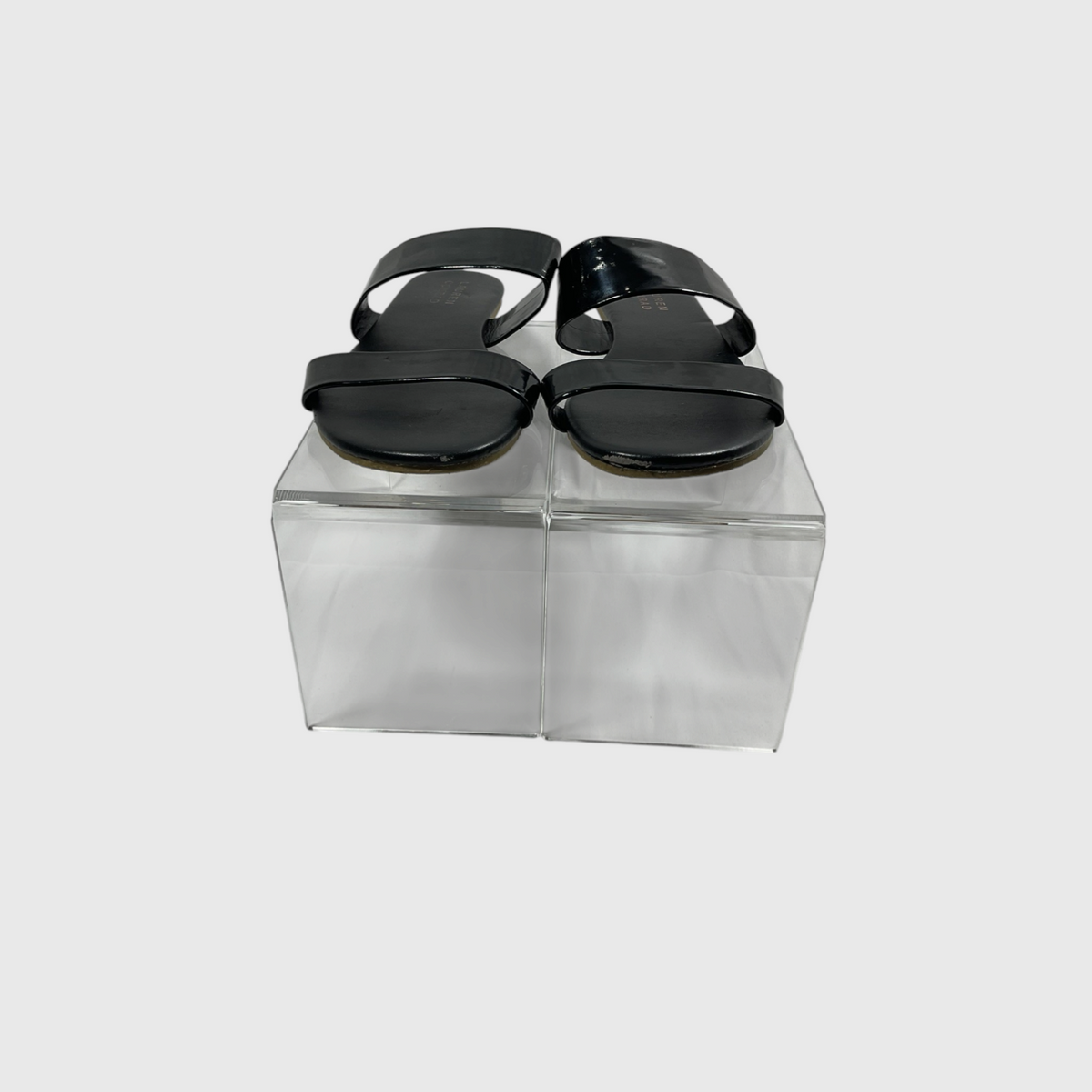 A pair of black patent leather slide sandals with wide double straps, displayed on a clear acrylic block against a light gray background.