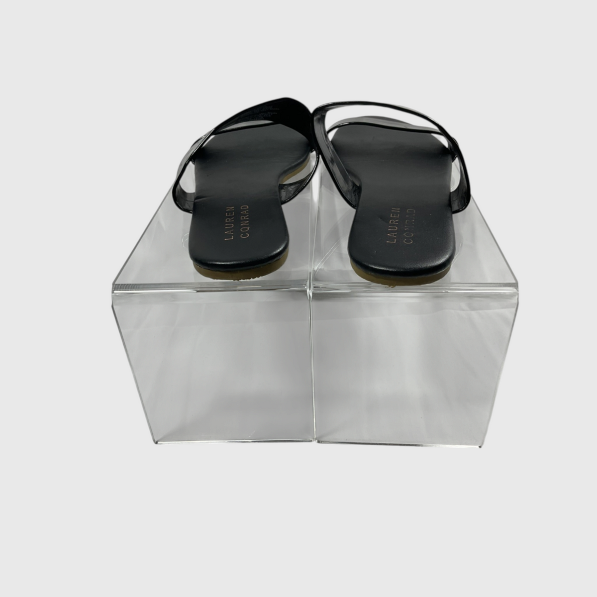 A pair of black flat thong sandals displayed on a clear acrylic block against a light gray background. The brand name 'LEATHER CRAFT' is printed on the insoles.