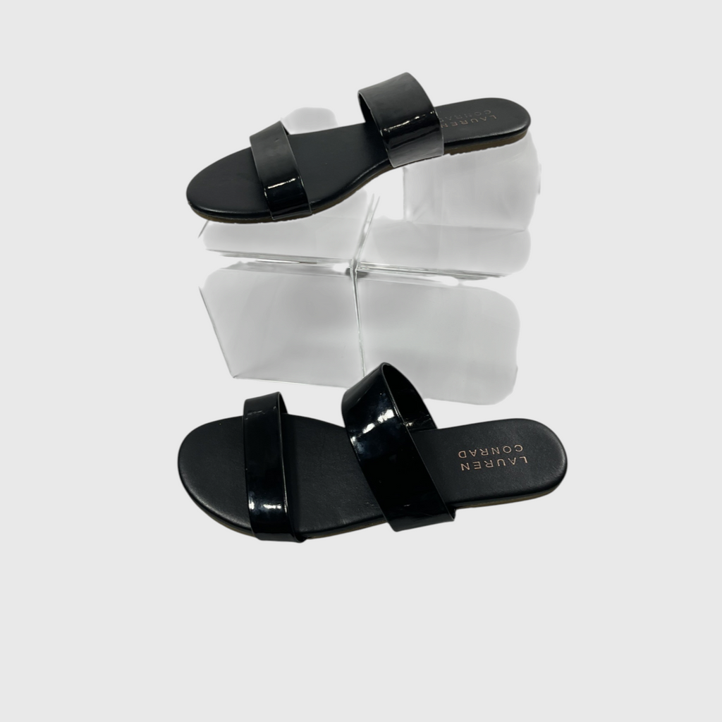 A pair of black patent leather slide sandals by Lauren Conrad, displayed on a clear acrylic block against a light gray background.