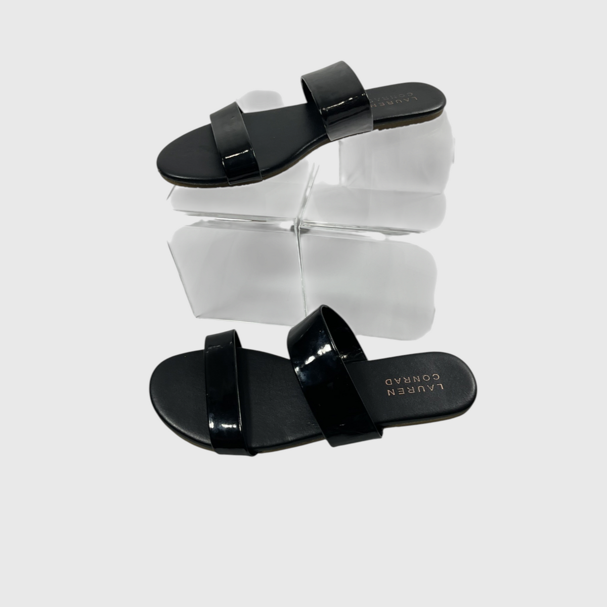 A pair of black patent leather slide sandals by Lauren Conrad, displayed on a clear acrylic block against a light gray background.