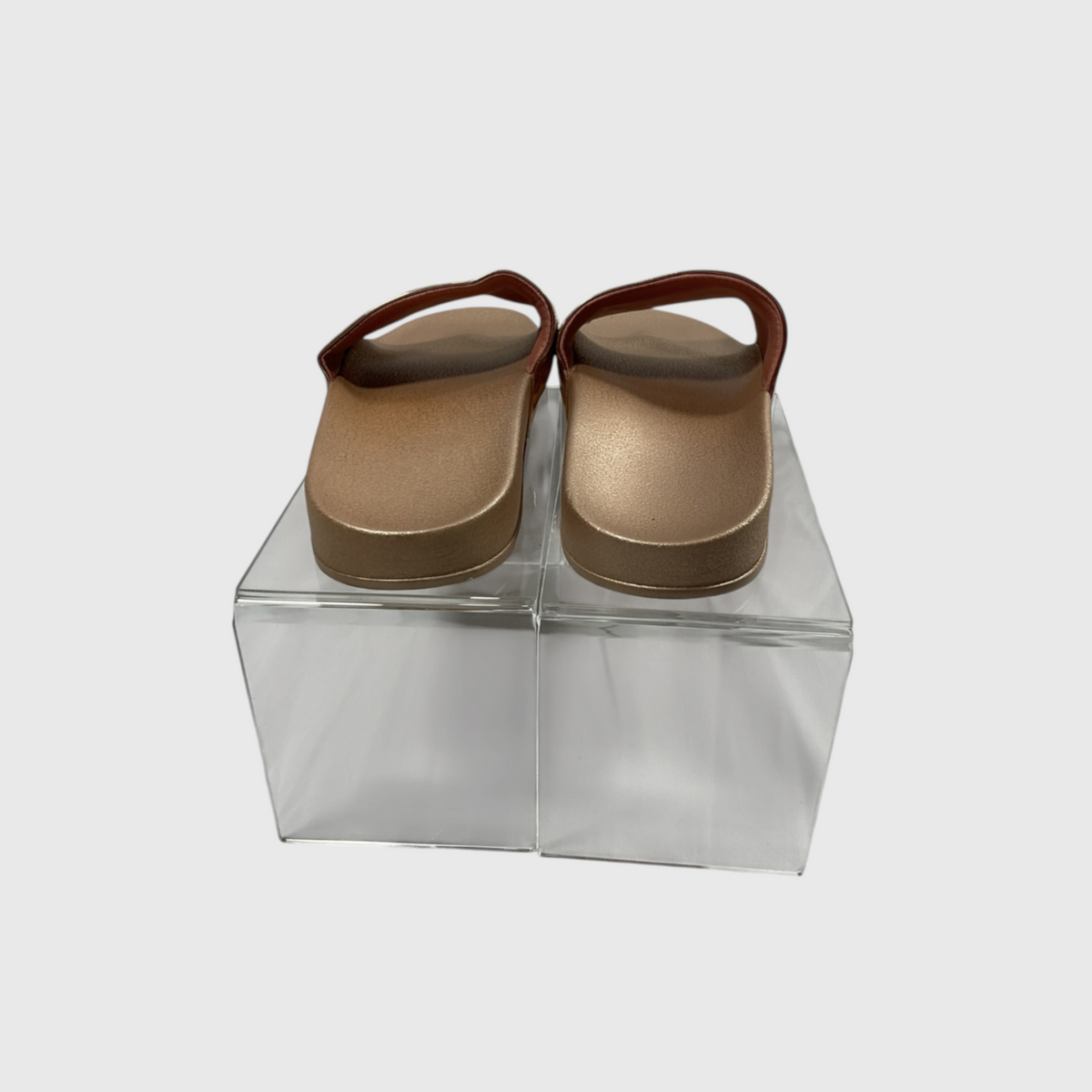 A pair of metallic gold clog-style sandals with brown straps, displayed on a clear acrylic block against a light gray background.