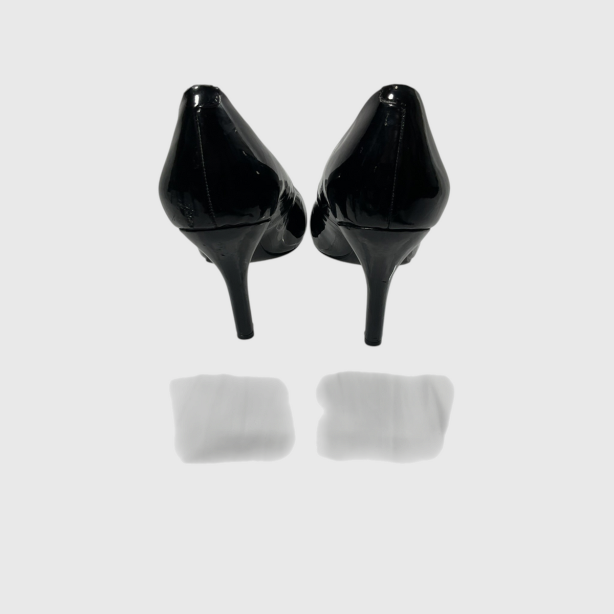 A pair of black patent leather high-heeled pumps shown from the back, positioned symmetrically on a light gray reflective surface.