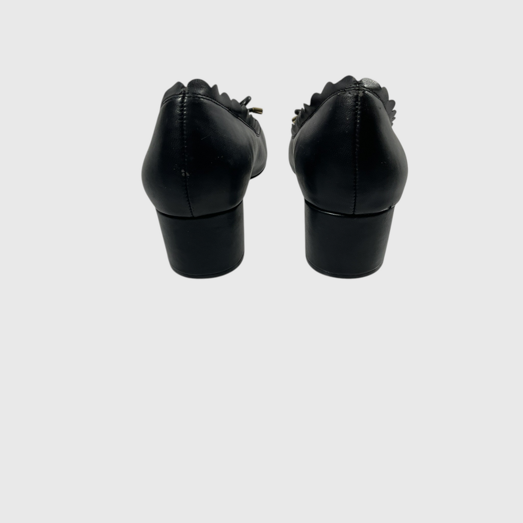 Black leather low-heeled pumps shown from the back, featuring a scalloped edge detail around the opening and a stacked block heel, set against a plain light gray background.