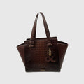 A dark brown tote bag with a crocodile-embossed front panel and smooth brown leather side panels and handles, featuring a gold-tone charm with an ampersand symbol and a small 'STUART' logo on the front, set against a light gray background.