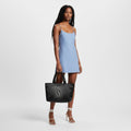 A model wearing a light blue slip dress and white strappy heels, holding a black speckled tote bag with a large embossed 'S' logo, set against a plain light gray studio background.