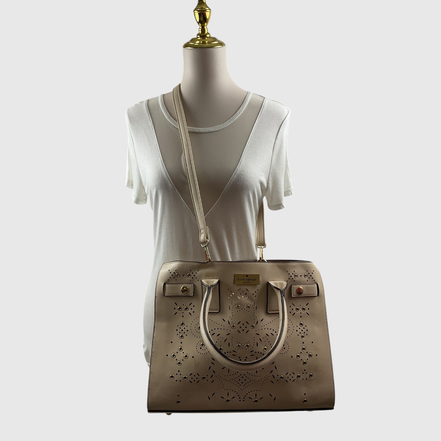 A beige perforated leather handbag with a shoulder strap, displayed on a white mannequin torso against a light gray background.
