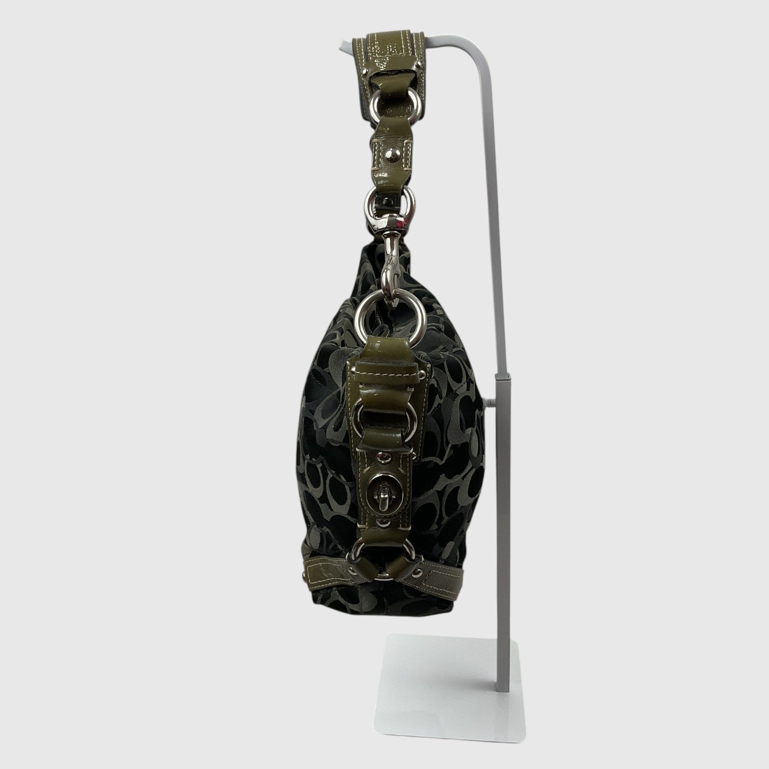 A small black and gray patterned handbag with olive green leather trim and silver-tone hardware, displayed on a white metal stand against a light gray background.