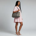 A woman modeling a gray canvas tote bag with dark brown leather trim and shoulder strap, worn crossbody over a light pink dress against a light gray studio background.