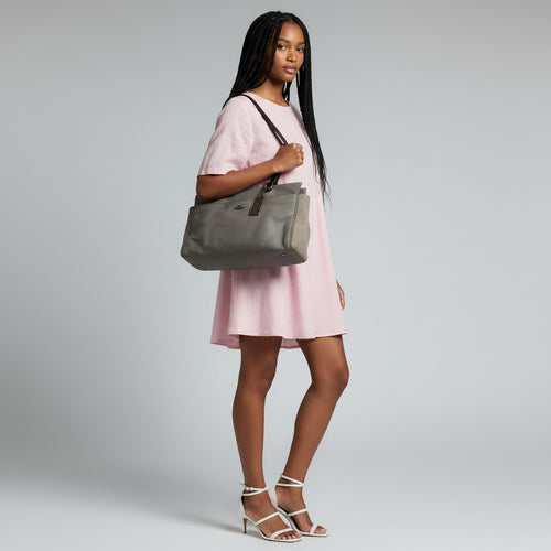 A woman modeling a gray canvas tote bag with dark brown leather trim and shoulder strap, worn crossbody over a light pink dress against a light gray studio background.